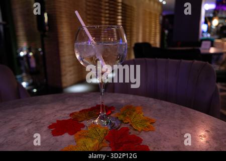 A refreshing drink with ice and a straw, surrounded by colourful autumn leaves on a marble table in a warm-lit setting. Stock Photo