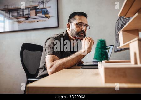 Male architect thoughtfully works at his desk on designing architectural models and plans, with the help of technology. Stock Photo