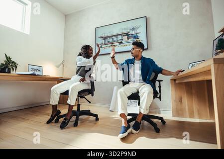 Two young architects, a woman and a man, celebrate a successful project with a high-five in a modern office setting, symbolizing teamwork, creativity, Stock Photo