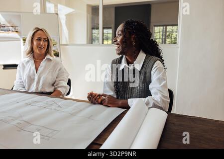 Two young female architects working together in an office, discussing blueprints and design ideas. The atmosphere is collaborative and friendly, refle Stock Photo