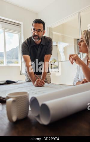 A male architect in his 40s collaborates with his team on blueprints at a desk. His colleagues assist while contributing ideas, showcasing professiona Stock Photo