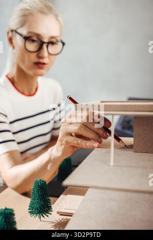 Middle-aged female architect wearing glasses working meticulously on a scale architectural model carefully. Stock Photo