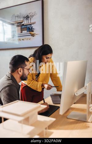 A young woman and a man collaborate at a desk in an architecture studio, reviewing designs on a computer. They are engaged in teamwork, crafting innov Stock Photo