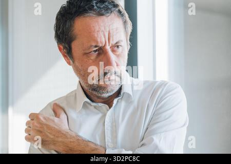 Unshaved mature businessman suffering from severe shoulder pain, holding arm in discomfort. Selective focus. Stock Photo