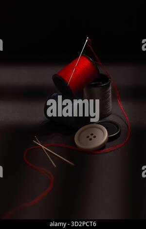 Black spool of thread, sewing needle and red thread arranged as a still life on a dark background, symbolizing tailoring. Stock Photo