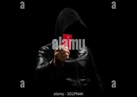 Close up to a thief faceless hacker hooded man holding a credit card with his right hand in the shadows Stock Photo