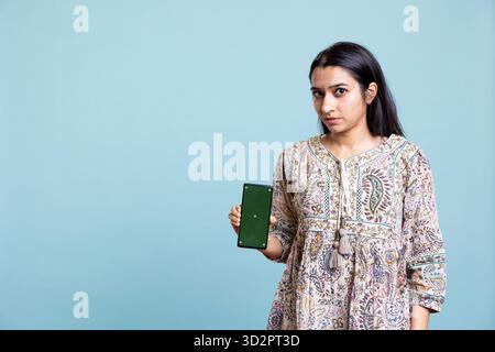 Happy indian woman presenting green screen on her smartphone, posing against the blue background. Joyous girl holding copy space device used for advertising brands in a commercial. Stock Photo