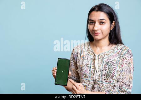 Happy indian woman presenting green screen on her smartphone, posing against the blue background. Joyous girl holding copy space device used for advertising brands in a commercial. Stock Photo