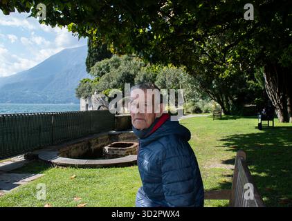 Elderly Man Sitting Alone on a Bench by Lake Como, Italy, Looking Sadly at the Camera on a Sunny Off-Season Day Stock Photo