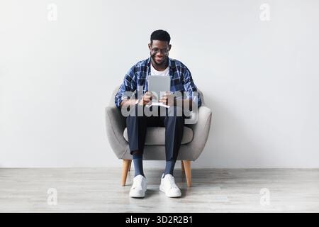 Young Black Man Enjoys Video Chat While Studying on Tablet in Cozy Armchair at Home Stock Photo