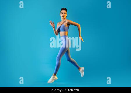 Fitness Female Jumping in Mid Air Against a Blue Background During a Workout Session Stock Photo