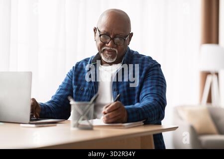 Mature African American Man Taking Notes While Using Laptop in Modern Office Environment Stock Photo