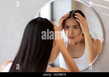 Worried Long-Haired Chinese Woman Checks Her Hair for Thinning in Bathroom Mirror Stock Photo