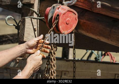 Determined worker hand pulling heavy metal chain on hoist for lifting large wood log. This shows manual labor and strength required in industry Stock Photo
