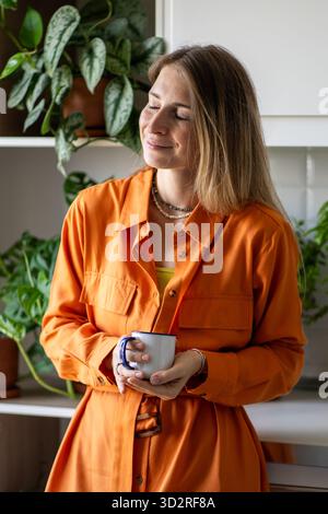 Positive scandinavian woman with mug of coffee in hands smiling with closed eyes standing in kitchen Stock Photo