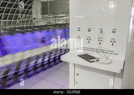 Charging phone on table of a charging station in airport terminal hall with moving row of luggage carts on a background. Stock Photo