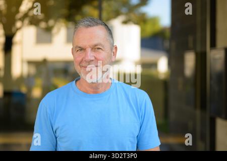 Calm and confident mature man with short gray hair and beard wearing a light blue shirt, smiling warmly while standing outside in soft daylight. Stock Photo