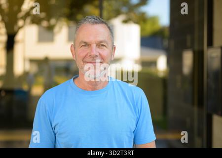 Relaxed middle-aged man with gray hair and beard wearing a blue shirt, smiling confidently while standing outdoors in bright natural light. Stock Photo