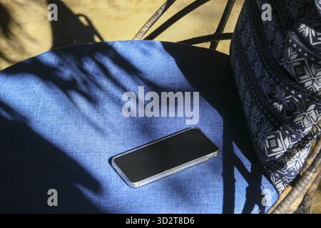 Quiet scene of mobile phone with blank screen resting on blue chair cushion in outdoor sunlight. smartphone creates dramatic, dark shadow on fabric Stock Photo