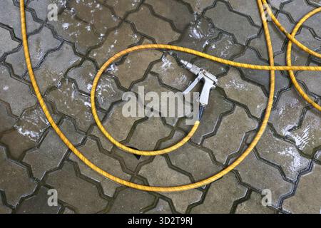 Yellow garden hose with spray nozzle resting on wet interlocking pavement. useful cleaning tool for outdoor watering or washing, on textured backgroun Stock Photo