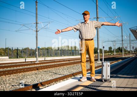 Angry senior man is standing at railway station. He is angry because train is late. Senior people traveling. Stock Photo