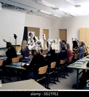 In the 1970s. A school class sits in the benches during a lesson with a female teacher. An overhead projector stands at the front, a common device in classrooms at the time that showed the transparent sheet with text and pictures placed on the device on the screen. Overhead projectors were widely used in education and business before the advent of computer-based projection. The teacher has asked a question and the student rises their hands to answer. Sweden 1970s Stock Photo