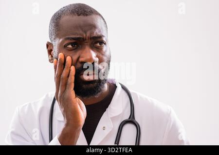 Portrait of a tired African American male doctor looking pensive, showing signs of emotional stress and exhaustion in a gray studio setting. Stock Photo