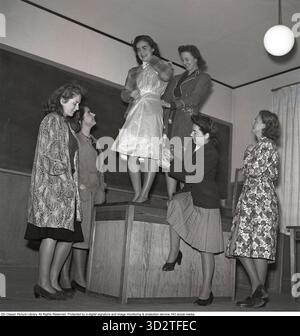 Students at a school for sewing and fashion design. In a sewing and fashion design school, students collaborate to ensure the correct length of a dress. One student is holding a skirt pattern while others assist in measuring the waist and dress length. In 1943, sewing and fashion design schools were pivotal in teaching practical skills for creating tailored clothing. Students learned techniques like pattern-making, measuring, and garment construction, often working together to master precision in designs, such as skirts and dresses, reflecting the era's emphasis on craftsmanship and resourcefu Stock Photo