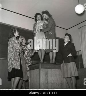 Students at a school for sewing and fashion design. In a sewing and fashion design school, students collaborate to ensure the correct length of a dress. One student is holding a skirt pattern while others assist in measuring the waist and dress length. In 1943, sewing and fashion design schools were pivotal in teaching practical skills for creating tailored clothing. Students learned techniques like pattern-making, measuring, and garment construction, often working together to master precision in designs, such as skirts and dresses, reflecting the era's emphasis on craftsmanship and resourcefu Stock Photo