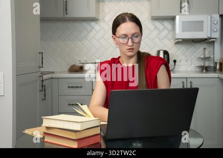 Focused female freelancer wearing glasses and a red shirt working from her home kitchen, using a laptop and taking notes in a notebook, with several b Stock Photo