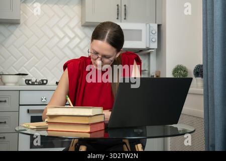 Focused woman in a stylish home office setting, writing notes in the notebook while working on a laptop. Modern kitchen in the background enhances the Stock Photo