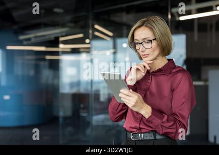 Businesswoman wearing glasses and a maroon shirt standing in a contemporary office, carefully reviewing data on a digital tablet with a focused and contemplative expression Stock Photo