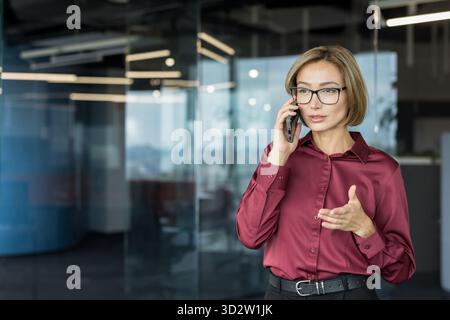 Professional businesswoman wearing glasses and a red shirt is having a phone conversation, actively gesturing and communicating in a modern corporate office environment Stock Photo