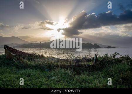 An Autumnal seascape at dawn with sun rays breaking through the clouds, and a broken wooden boat in the foreground. Stock Photo