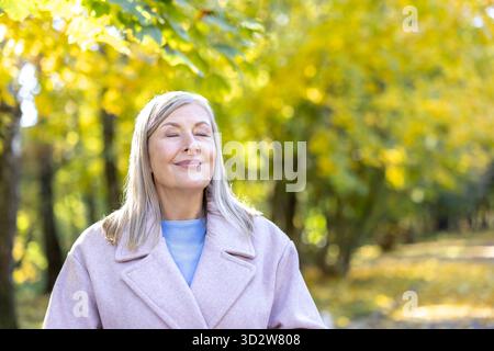 Senior woman relaxing with eyes closed and a gentle smile, feeling the sun on her face in an autumn park with blurred yellow foliage in the background Stock Photo