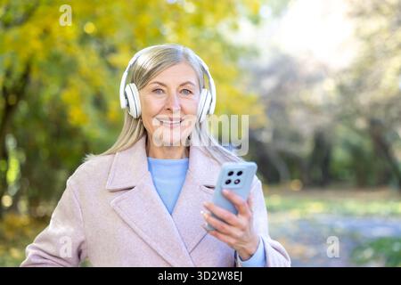 Senior woman smiling and listening to music through headphones, holding a smartphone while relaxing outdoors in an autumn park, embracing technology and leisure Stock Photo