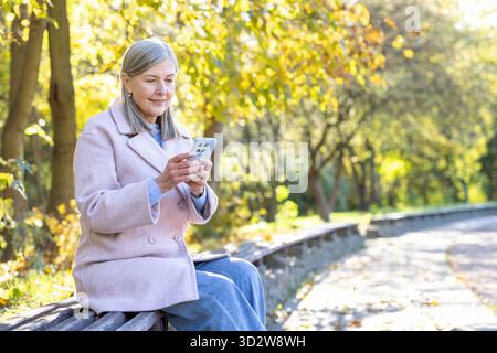 Senior woman relaxing on a park bench while engaging with her smartphone, enjoying digital connectivity and serene leisure surrounded by the vibrant colors of autumn foliage Stock Photo