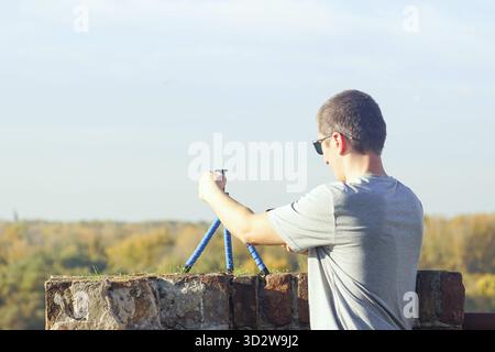 Person films himself using a smartphone mounted on a compact travel-friendly tripod in a location with a panoramic view. Symbol of modern tourism. Stock Photo
