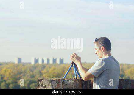 Man filming cityscape panorama with smartphone mounted on tripod at viewpoint. Concept of timelapse video recording and travel content production. Stock Photo