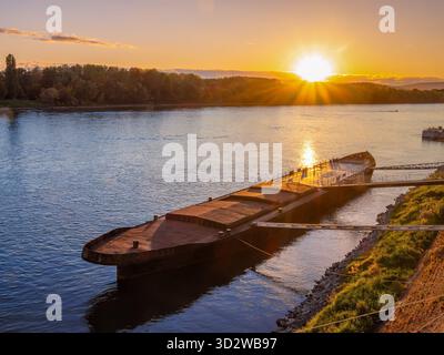 A barge docked on a Danube river in Bratislava during sunset, with the sun setting on the horizon and reflecting on the water Stock Photo