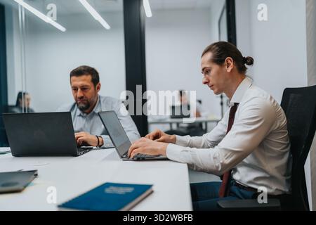 Professional office scene with two coworkers working on laptops and collaborating Stock Photo