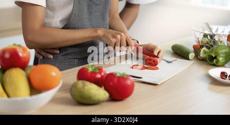 In a bright kitchen, a couple is engaged in cooking, slicing fresh tomatoes while sharing smiles and laughter Stock Photo
