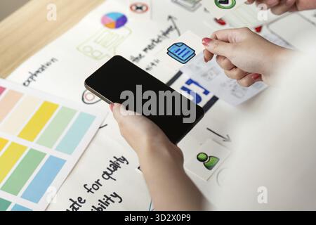 Creative process in UX and UI design with hands holding a smartphone and design elements on a table. Focus on user experience and interface Stock Photo