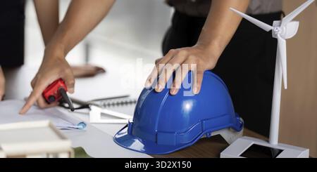 An engineer using tools and a blue hard hat while planning innovative sustainable energy solutions with models and blueprints on a table Stock Photo