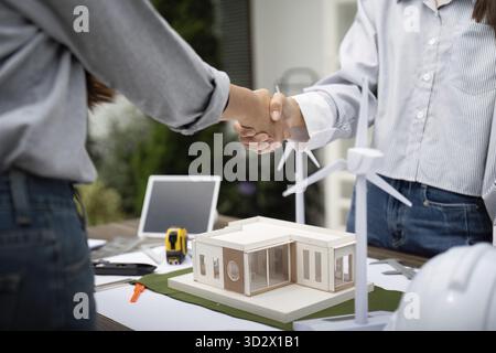 Two sustainability engineers shaking hands as a sign of agreement on their project, with architectural models and blueprints on the table outdoors Stock Photo