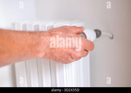 man regulates the temperature in the house with a thermostat on the white radiator, close-up male hand, concept of saving heat, beginning of the heati Stock Photo