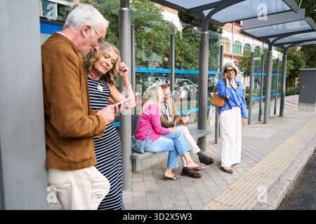 Group of senior friends and couples interacting with smartphones at a city bus shelter, waiting for public transportation Stock Photo