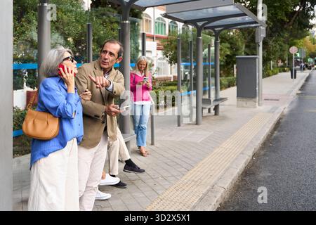 Senior couple standing at a bus stop, man pointing away, woman talking on phone, another woman using phone Stock Photo