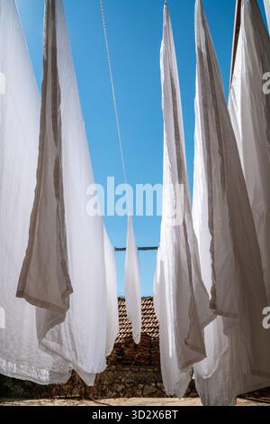 Washing day under bright blue sky. Crisp white sheets billow gently in the sun, creating dramatic lines against rustic terracotta roof and stone wall Stock Photo
