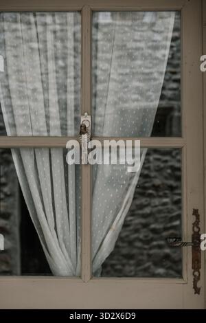 Vintage window with sheer dotted curtains and a small decorative bird ornament hanging on the wooden frame. Stock Photo